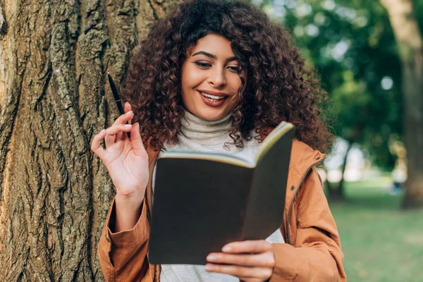 Foco seletivo da jovem mulher segurando caneta e olhando para notebook perto da árvore no parque — Fotografia de Stock