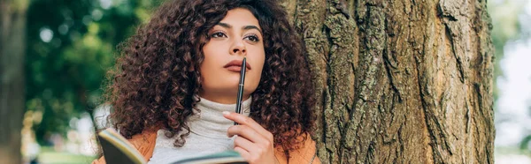 Horizontal crop of dreamy woman with pen and notebook looking away near tree — Stock Photo