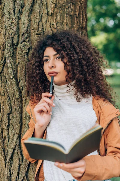 Selective focus of pensive woman in autumn outfit holding pen and notebook in park — Stock Photo