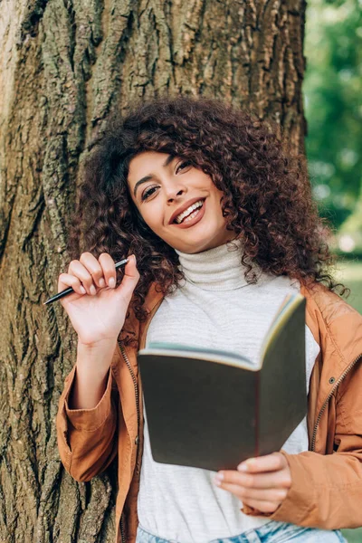 Selektiver Fokus der lockigen Frau mit Notizbuch und Stift, die in Baumnähe in die Kamera blickt — Stockfoto