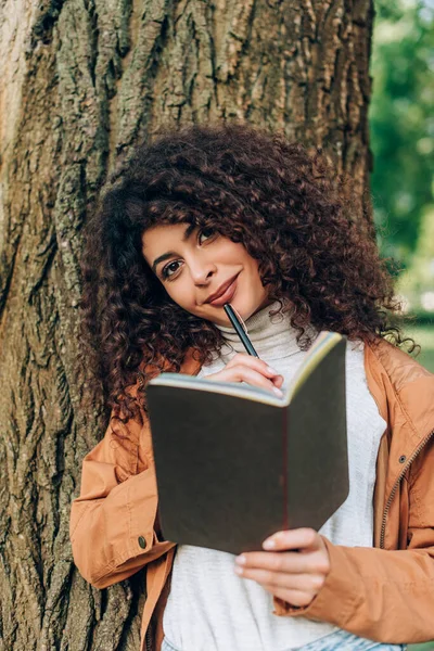 Enfoque selectivo de la mujer joven en impermeable que sostiene la pluma y el cuaderno mientras mira la cámara cerca del árbol - foto de stock