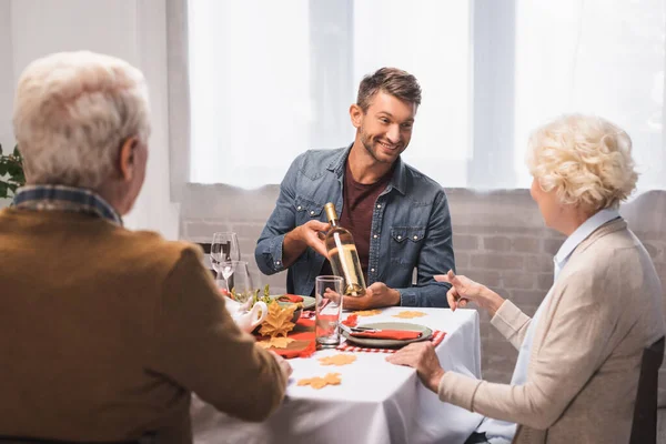 Seniorin zeigt beim Erntedankessen mit Finger auf Weinflasche in den Händen ihres fröhlichen Sohnes — Stockfoto