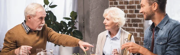 Горизонтальное изображение пожилого человека, указывающего пальцем на бутылку вина в руках сына во время праздничного ужина — стоковое фото