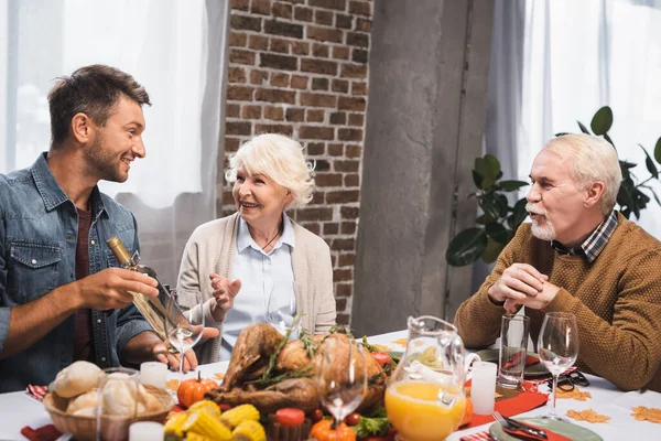 Joyful man holding bottle of white wine while celebrating thanksgiving with senior parents — Stock Photo