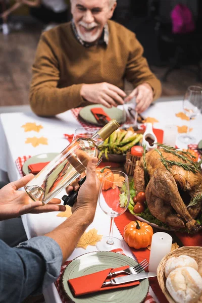 Enfoque selectivo del hombre sosteniendo botella de vino blanco mientras está sentado en la mesa servida con cena de acción de gracias - foto de stock