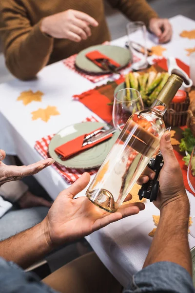 Cropped view of man holding bottle of white wine during thanksgiving dinner with family — Stock Photo