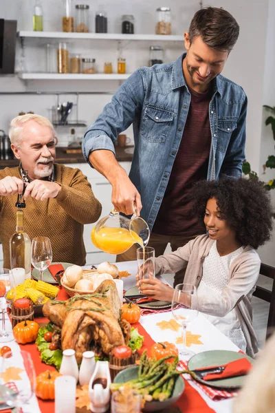 Enfoque selectivo de los hombres que abren la botella de vino blanco y vierten jugo de naranja mientras celebran la acción de gracias con la familia multicultural - foto de stock