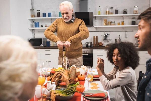 Hombre mayor abriendo botella de vino blanco durante la cena de acción de gracias con la familia multiétnica - foto de stock