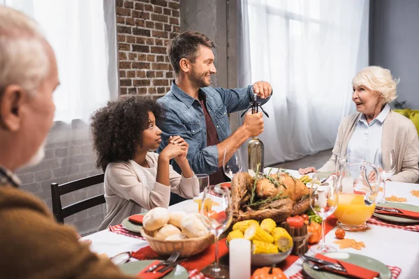 Man opening bottle of white wine during thanksgiving dinner with multicultural family — Stock Photo