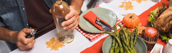 Cropped view of man holding bottle of white wine and corkscrew at table served with thanksgiving dinner, horizontal image — Stock Photo