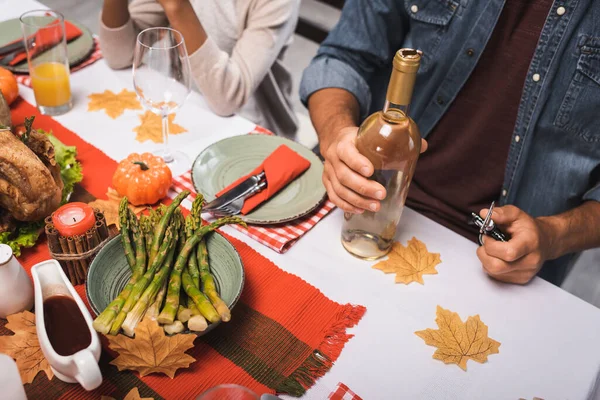 Vista ritagliata di uomo in possesso di bottiglia di vino mentre seduto vicino ragazza afro-americana durante la cena del Ringraziamento — Foto stock