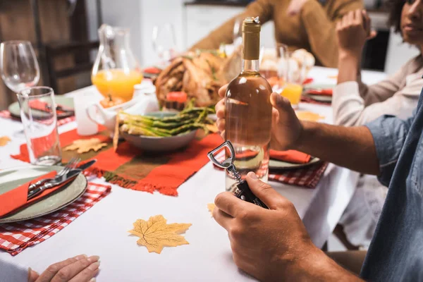 Cropped view of man holding bottle of white wine and corkscrew during thanksgiving dinner with multicultural family — Stock Photo
