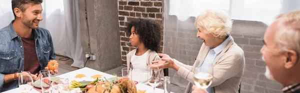 Tiro panorâmico de mulher excitada gesticulando durante o jantar de ação de graças com a família multicultural — Fotografia de Stock