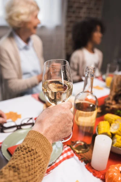 Hombre mayor sosteniendo una copa de vino blanco mientras celebra el Día de Acción de Gracias con la familia multicultural - foto de stock