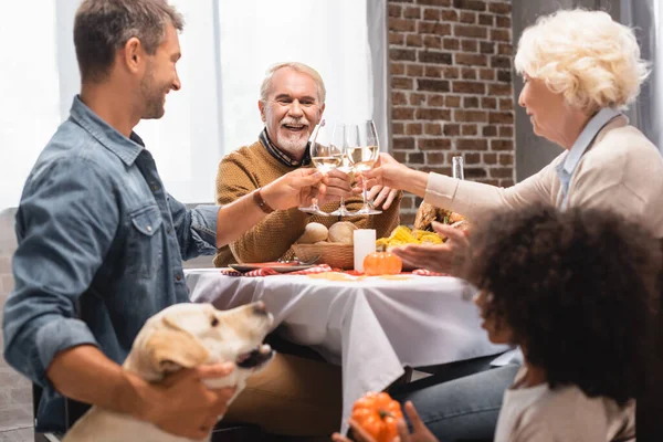 Enfoque selectivo de chica afroamericana y golden retriever cerca de la familia tintineo copas de vino durante la cena de acción de gracias - foto de stock