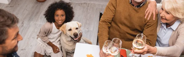 Selective focus of african american girl and golden retriever near family clinking wine glasses on thanksgiving day, panoramic shot — Stock Photo