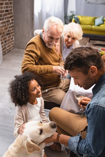 Homme caressant golden retriever et tenant un verre de vin blanc pendant le dîner de Thanksgiving avec une famille multiculturelle — Photo de stock