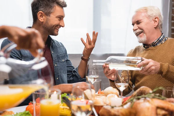 Joyful man gesturing while talking to father pouring white wine during thanksgiving dinner — Stock Photo