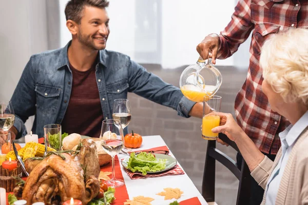 Alegre hombre sentado en la mesa con la cena de acción de gracias mientras que la mujer afroamericana verter jugo de naranja - foto de stock