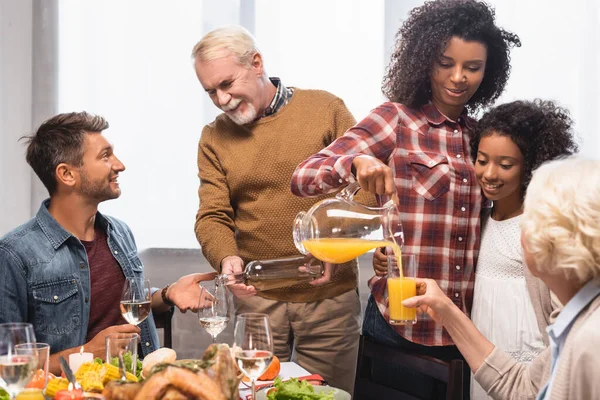 Mujer afroamericana y hombre mayor vertiendo jugo de naranja y vino blanco durante la cena de acción de gracias con la familia multicultural - foto de stock