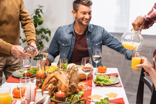 Homem animado sentado à mesa servido com jantar de ação de graças, enquanto parentes multiculturais derramando bebidas em copos — Fotografia de Stock