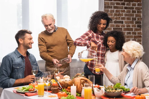 Freudige afrikanisch-amerikanische Frau und älterer Mann schenken Orangensaft und Weißwein während des Dankeschön-Abendessens mit multiethnischen Familie ein — Stockfoto