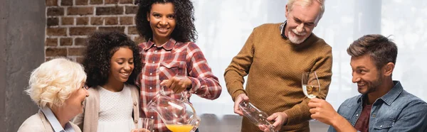 Panoramic shot of senior man and african american woman pouring orange juice and white wine during thanksgiving dinner with multicultural family — Stock Photo