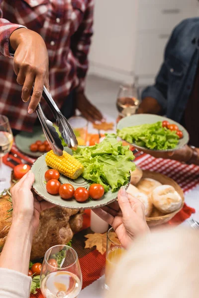 Partial view of senior woman holding plate with corn, lettuce and cherry tomatoes during thanksgiving dinner with multiethnic family — Stock Photo