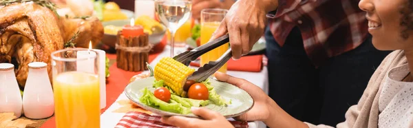 Vista recortada de la madre poniendo maíz en el plato para la hija afroamericana durante la cena de acción de gracias, tiro panorámico - foto de stock