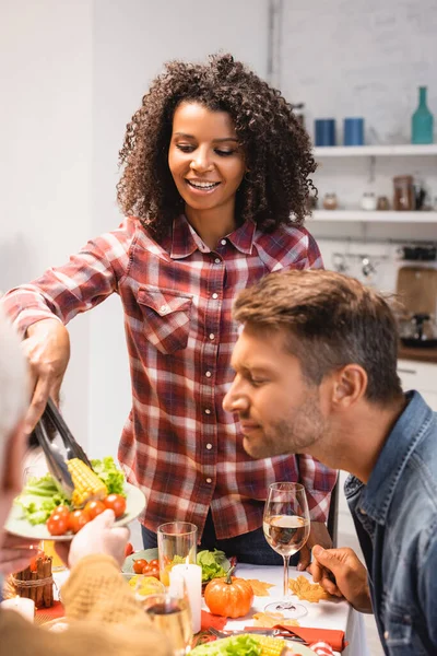 Enfoque selectivo del hombre con los ojos cerrados cerca de la esposa afroamericana tomando maíz durante la cena de acción de gracias - foto de stock