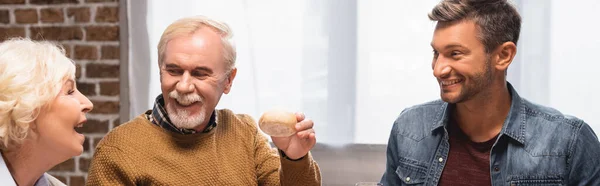 Panoramaaufnahme eines älteren Mannes, der am Erntedankfest neben freudiger Frau und Sohn ein Brötchen hält — Stockfoto