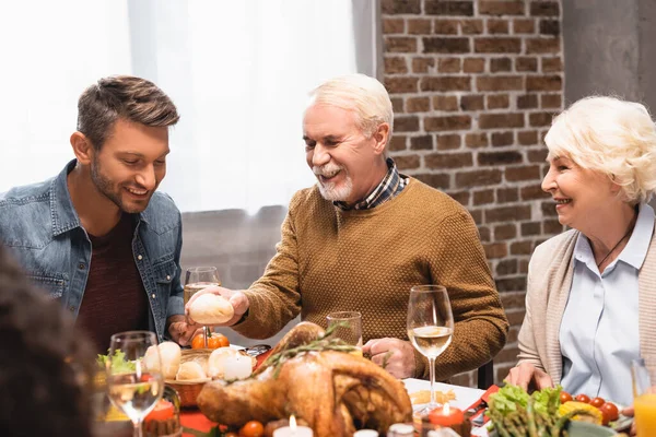 Fröhlicher Senior hält Brötchen, während er Erntedankfest mit Familie feiert — Stockfoto