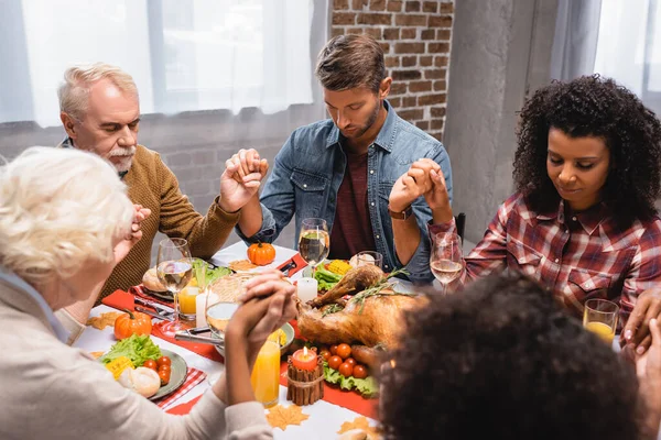 Foyer sélectif de la famille multiethnique avec les yeux fermés tenant la main pendant le dîner pendant les vacances de thanksgiving — Photo de stock