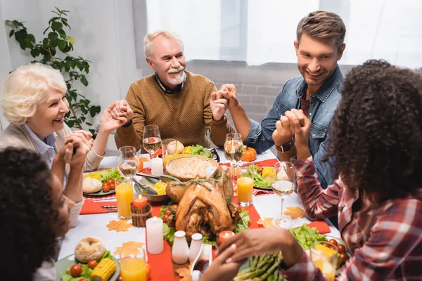 Familia multiétnica alegre tomados de la mano durante la cena en las vacaciones de acción de gracias - foto de stock