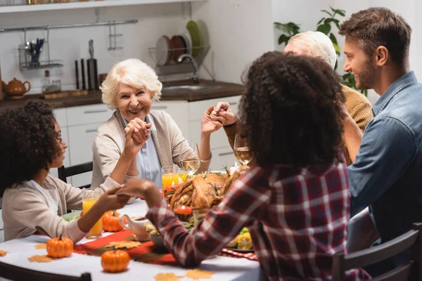 Enfoque selectivo de la familia multicultural alegre tomados de la mano durante la cena en las vacaciones de acción de gracias - foto de stock