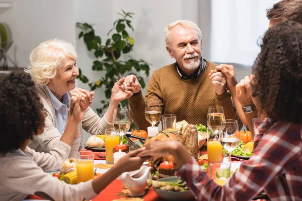 Familia multiétnica cogida de la mano cerca de la cena en vacaciones de acción de gracias - foto de stock