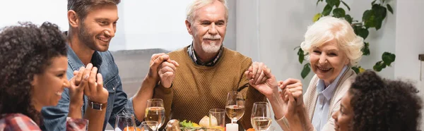 Panoramic shot of pleased multiethnic family holding hands on thanksgiving — Stock Photo