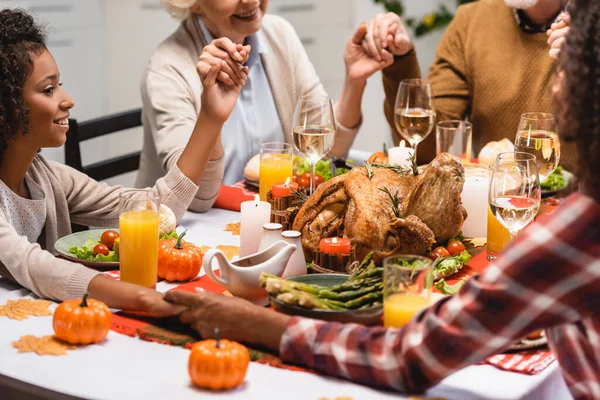 Parcial da família multiétnica de mãos dadas perto da mesa com jantar de ação de graças — Fotografia de Stock