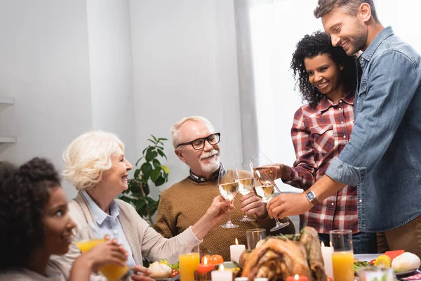 Enfoque selectivo de alegre familia multiétnica tintineo vasos de vino blanco durante la cena de Acción de Gracias — Stock Photo