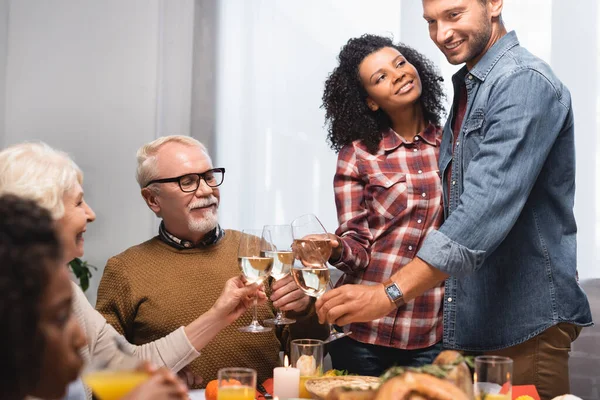 Selektiver Fokus multikultureller Familie, die beim Erntedankfest mit Weißweingläsern klingelt — Stockfoto