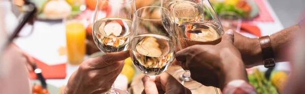 Cropped view of multiethnic family clinking glasses of white wine during Thanksgiving dinner, horizontal image — Stock Photo