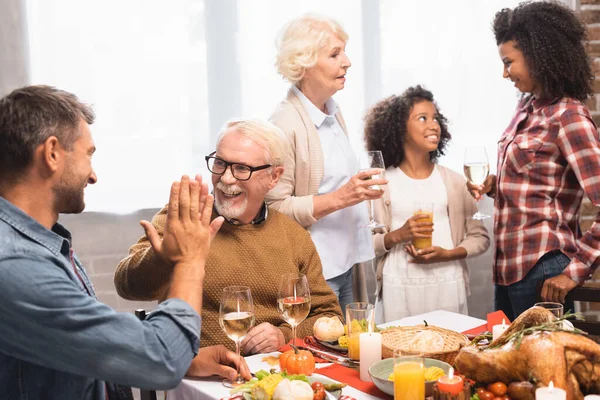 Hombres alegres dando cinco altos durante la cena de acción de gracias con la familia multicultural - foto de stock