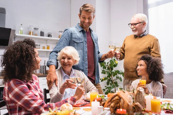 Alegre familia multicultural celebrando el día de acción de gracias en la mesa servida con deliciosa cena - foto de stock