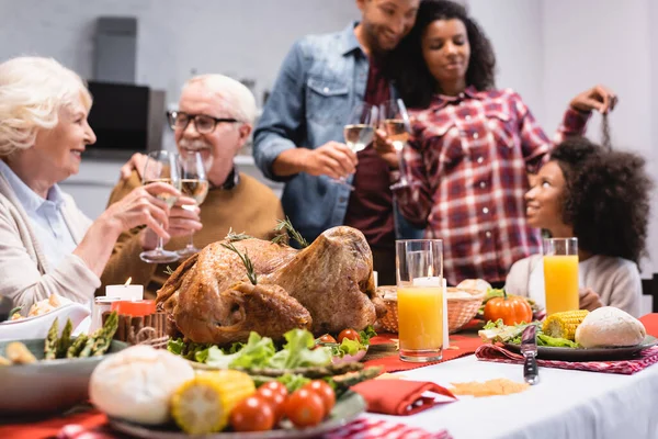 Enfoque selectivo del sabroso pavo en la mesa cerca de la familia multiétnica con copas de vino durante la celebración de Acción de Gracias - foto de stock