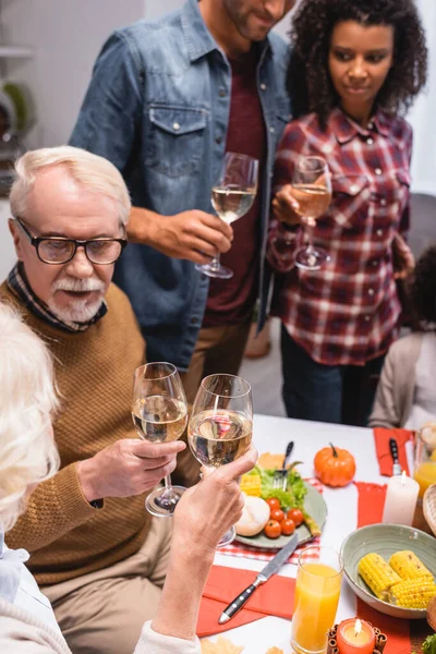 Foco seletivo da mulher idosa batendo vinho com o homem durante a celebração de ação de graças com a família multiétnica — Fotografia de Stock