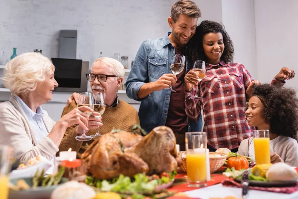 Enfoque selectivo de la familia multicultural con copas de vino sentadas a la mesa con comida sabrosa durante el Día de Acción de Gracias - foto de stock