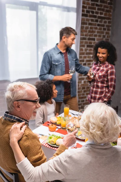 Focus selettivo della donna anziana che abbraccia il marito con un bicchiere di vino vicino alla famiglia multietnica durante il Ringraziamento — Foto stock