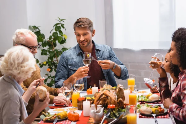 Enfoque selectivo del hombre sosteniendo el vino cerca de la familia multiétnica durante la cena de acción de gracias en casa - foto de stock