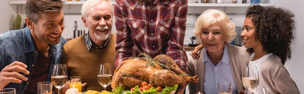 Cultivo panorámico de mujer sosteniendo pavo mientras celebra el Día de Acción de Gracias con una familia multiétnica - foto de stock