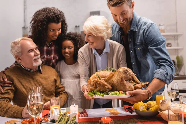 Selective focus of elderly woman holding tray with turkey near multicultural family during thanksgiving — Stock Photo